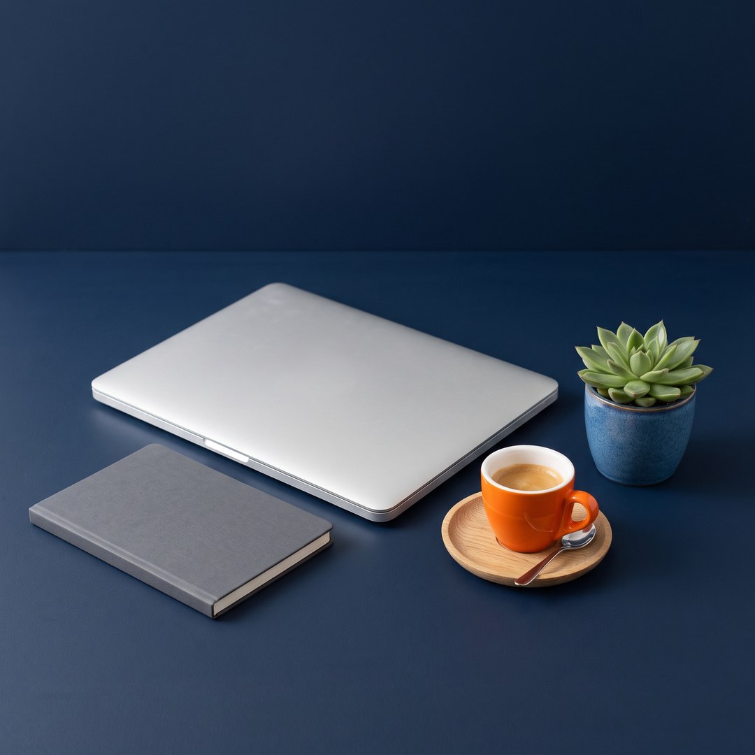 Een dichtgeklapte laptop, een grijs notitieboek, een oranje espressokopje op een houten schoteltje en een kleine vetplant op een donkerblauw bureau.